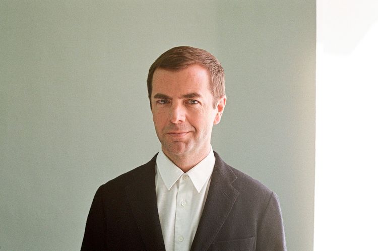 Ein Mann mit kurzem braunem Haar trägt ein weißes Hemd und ein dunkles Sakko. Er schaut direkt in die Kamera, der Hintergrund ist neutral mit weichen Lichtreflexionen.