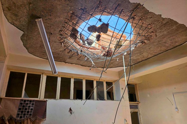 Das Bild zeigt einen stark beschädigten Raum mit einem großen Loch in der Betondecke, durch das Himmel und Ziegelteile sichtbar sind. Herabhängende Stahlstangen und Kabel zeugen von der Zerstörung. Unter der Decke sind Fensterreihen und eine beschädigte Wand sichtbar.