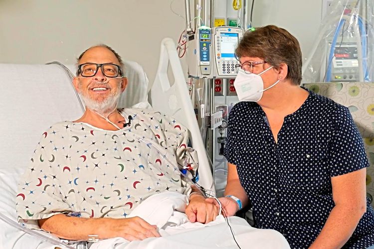 Der Mann liegt lächelnd im Krankenbett, seine Frau sitzt daneben und hält seine Hand.