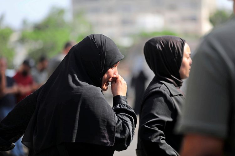 Zwei trauernde Frauen in schwarzer Kleidung, eine mit einem Schleier, die andere mit einem Hijab, drückt sich die Hand ins Gesicht. Sie stehen im Freien, im Hintergrund sind weitere Personen unscharf zu sehen.