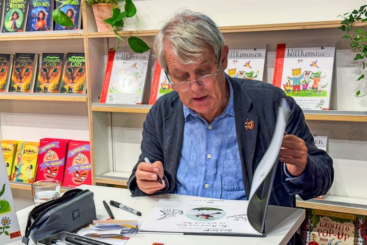 Ein Mann sitzt an einem Tisch, signiert und zeichnet in ein Buch. Auf dem Tisch liegen Stifte, ein Federmäppchen und Papier. Im Hintergrund sind Bücher und Dekorationen in einem Bücherregal zu sehen.