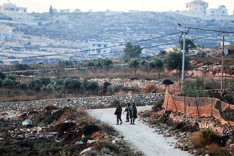 Drei Soldaten gehen auf einem unbefestigten Weg in einer ländlichen Landschaft nahe Qusra im israelisch besetzten Westjordanland. Im Hintergrund sind Häuser, Olivenbäume und Hügel zu sehen. Stromleitungen verlaufen über die Szenerie.
