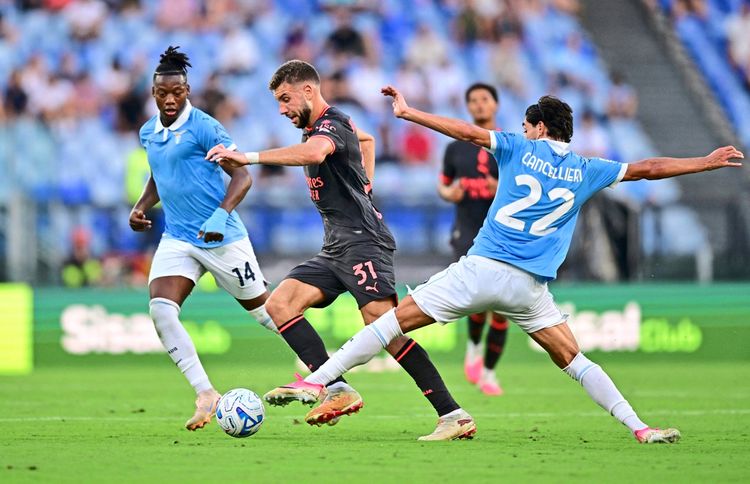Fußballspiel in der Serie A zwischen Lazio und AC Mailand im Stadio Olimpico in Rom. Ein Spieler von AC Mailand (in schwarzem Trikot) dribbelt den Ball, während zwei Lazio-Spieler (in hellblauen Trikots) versuchen, den Ball zu erobern. Im Hintergrund unscharfe Zuschauertribünen.