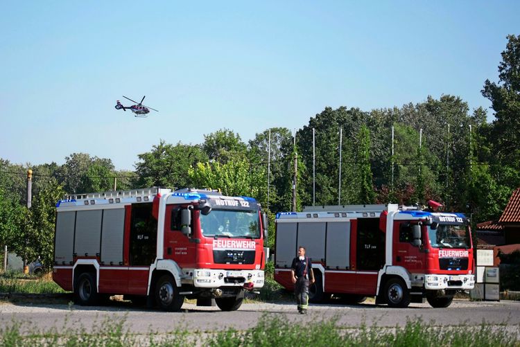 Zwei Feuerwehr-Einsatzfahrzeuge stehen nebeneinander auf einer Straße, während ein Feuerwehrhelfer daneben läuft. Im Hintergrund ist ein Helikopter in der Luft zu sehen, umgeben von Bäumen.