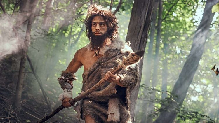 Foto eines als Neandertaler hergerichteten Mannes im Wald, er trägt lange, verfilzte braune Haare und Bart, einen Speer und Felle.