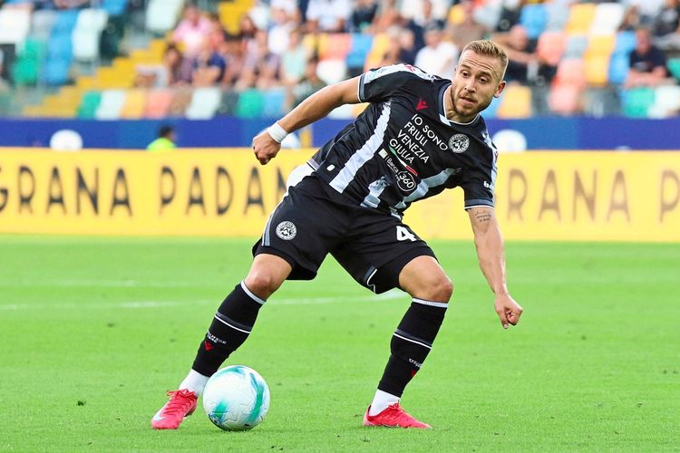 Ein Fußballspieler von Udinese, in Schwarz-Weiß gestreiftem Trikot, kontrolliert den Ball während eines Spiels im Bluenergy-Stadion in Udine. Im Hintergrund Zuschauer und Werbebanden.