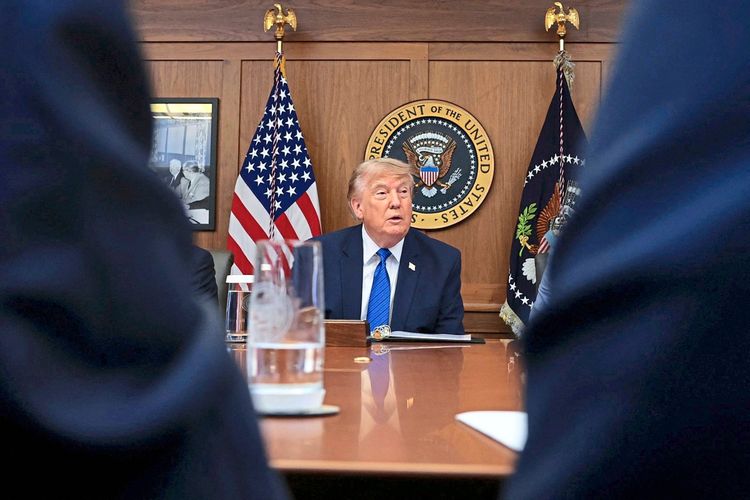 Ein Mann sitzt an einem Tisch vor zwei Flaggen der USA und dem Siegel des Präsidenten der Vereinigten Staaten. Im Hintergrund ist eine Holzvertäfelung sowie ein gerahmtes Bild sichtbar. Im Vordergrund sind unscharf Gläser und Personen angedeutet.