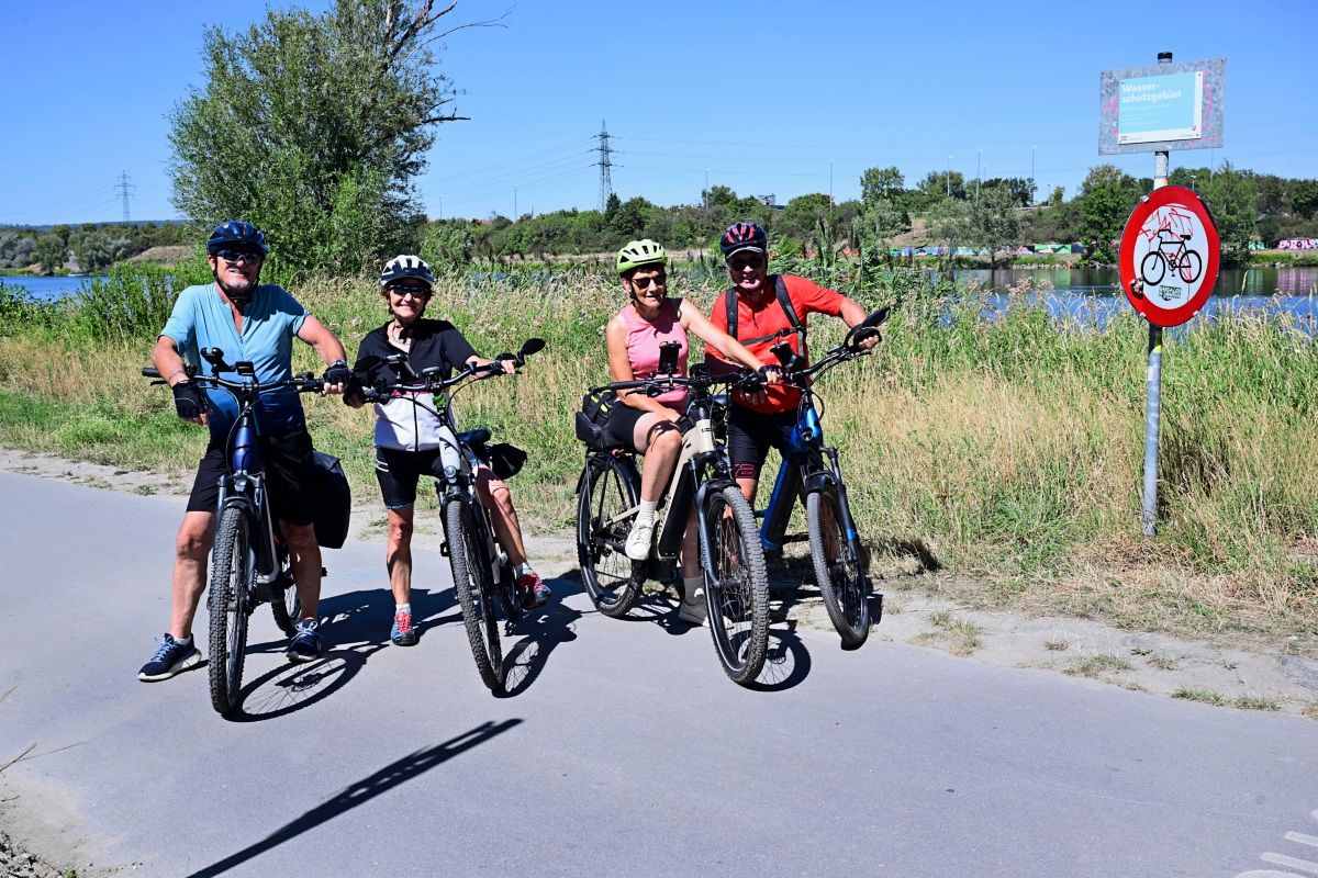 Vier Personen stehen mit ihren Fahrrädern auf einem asphaltierten Weg in einer sommerlichen Landschaft. Im Hintergrund sind Bäume, ein See und ein Schild mit einem Fahrrad-Symbol zu sehen. Die Gruppe trägt sportliche Kleidung und Fahrradhelme.