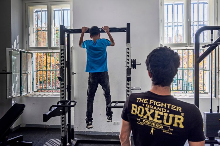 Ein Jugendlicher macht Klimmzüge an einem Fitnessgerät in einem hellen Raum mit hohen Fenstern. Ein weiterer Jugendlicher im Vordergrund beobachtet ihn.