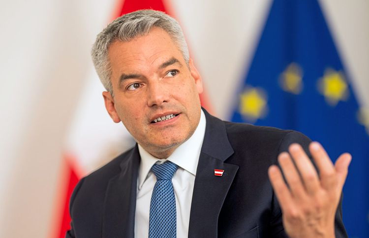 Bundeskanzler Karl Nehammer trägt einen Anzug mit blauer Krawatte. Im Hintergrund sind die österreichische und EU-Flagge zu sehen.