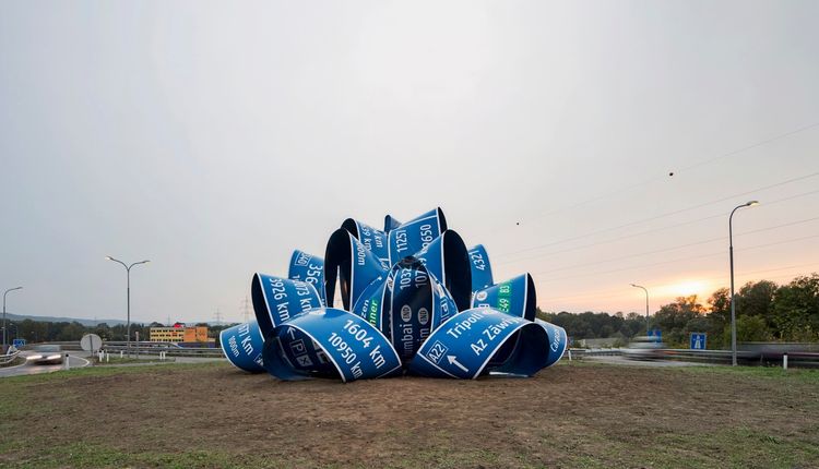 Eine große Skulptur aus blauen Verkehrsschildern, die zu einer verschlungenen, unregelmäßigen Form zusammengerollt sind, steht auf einem Kreisverkehr. Die Schilder zeigen Ortsnamen und Entfernungsangaben. Im Hintergrund sind Straßenlaternen, vorbeifahrende Autos und Bäume zu sehen.