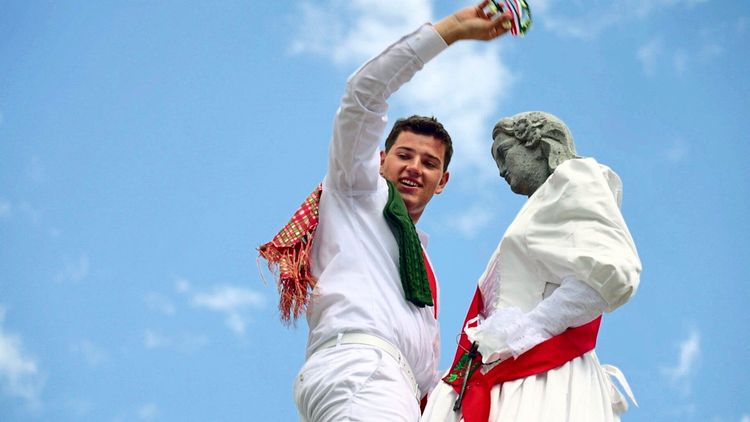 Ein Mann in traditioneller Kleidung mit weißem Hemd und Hose sowie buntem Halstuch und Tuch über der Schulter steht neben einer bekleideten Statue. Er hält einen bunten Kranz in der Hand. Der Hintergrund zeigt einen blauen Himmel.