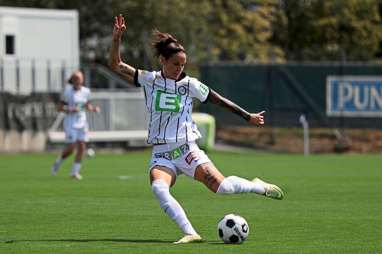 Eine Fußballspielerin im gestreiften Trikot von SK Sturm Graz Frauen ist mitten in einer Schussbewegung auf dem Spielfeld. Im Hintergrund ist eine weitere Spielerin sowie Teile der Tribüne und Werbebanner zu sehen.