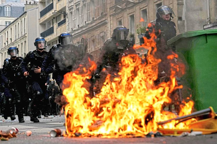 Eine Gruppe von Polizeiangehörigen in Schutzmontur schreitet energisch voran, während im Vordergrund ein brennender Mülleimer auf einer Straße in Paris steht.