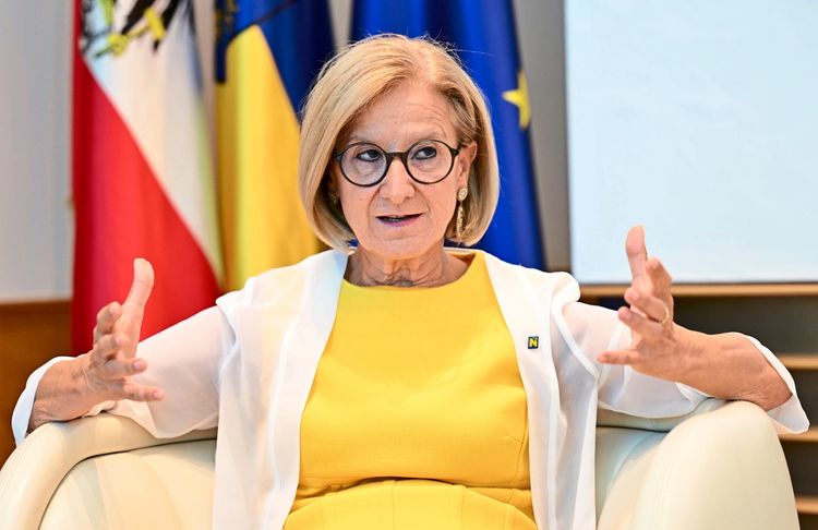 Eine Frau mit blonden Haaren und Brille sitzt in einem Sessel. Sie trägt ein gelbes Kleid mit einer weißen Jacke darüber und gestikuliert mit den Händen. Im Hintergrund sind unscharf Flaggen erkennbar, darunter die österreichische Flagge.