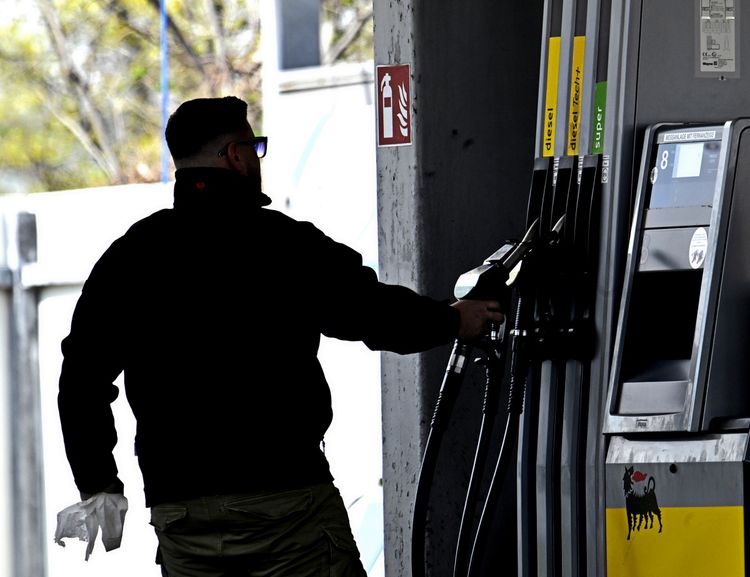 Ein Mann tankt an einer Zapfsäule an einer Tankstelle. Er trägt dunkle Kleidung und hält ein Zapfventil in der Hand. Im Hintergrund sind die Tanksäulen mit Beschriftungen wie 