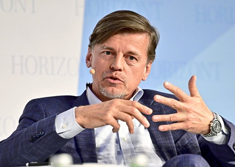 Ein Mann in einem blauen Anzug spricht und gestikuliert während einer Diskussionsveranstaltung. Er trägt ein Mikrofon am Ohr. Im Hintergrund ist unscharf der Schriftzug 