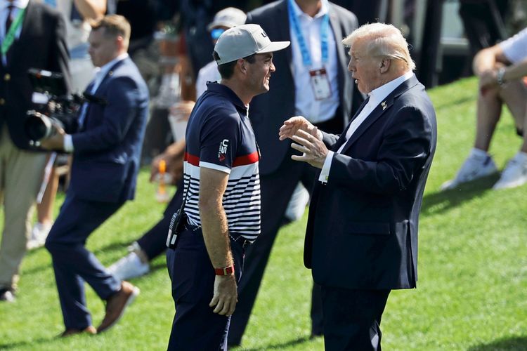 Zwei Männer stehen auf einem Golfplatz und unterhalten sich. Eine der Personen trägt ein gestreiftes Team-Poloshirt und eine Baseballkappe, während die andere einen dunklen Anzug trägt. Im Hintergrund sind weitere Personen, darunter ein Kameramann, zu sehen.