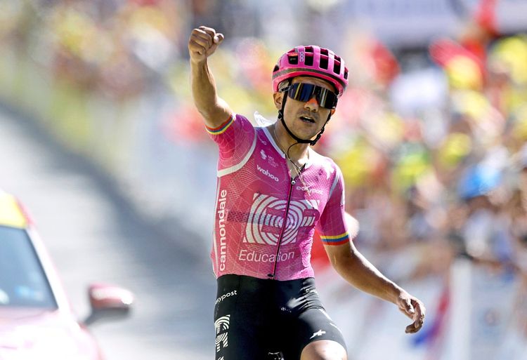 Ein Radfahrer im pinkfarbenen Trikot von EF Education-EasyPost jubelt mit erhobener Faust über den Sieg bei der 18. Etappe der Tour de France. Im Hintergrund sind unscharf Zuschauer mit bunten Farben zu sehen.