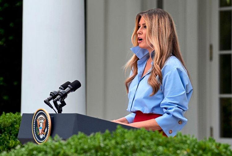 Eine Frau steht im Rosengarten des Weißen Hauses in Washington, D.C., hinter einem Rednerpult mit dem Siegel des Präsidenten der Vereinigten Staaten. Sie hält eine Rede und trägt eine blaue Bluse und einen roten Rock. Im Hintergrund sind ein Teil einer Säule und Fenster des Gebäudes zu sehen.
