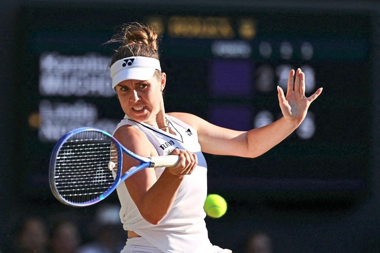 Tennisspielerin während eines Spiels in Aktion, hält einen Schläger und schlägt einen Ball zurück. Sie trägt ein weißes Sportoutfit und ein Stirnband, der Hintergrund zeigt unscharfe Anzeigetafeln.
