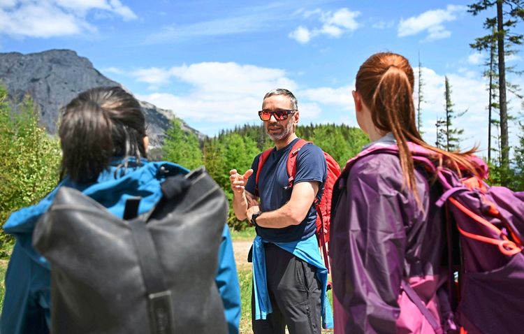 Eine Gruppe von drei Personen auf einer Wanderung in einer bergigen Landschaft. Im Vordergrund zwei Personen von hinten, beide mit Rucksäcken, im Hintergrund ein Mann mit Sonnenbrille, rotem Rucksack und in sportlicher Kleidung, der gestikuliert. Umgeben von Bäumen, blauen Himmel und Bergen.