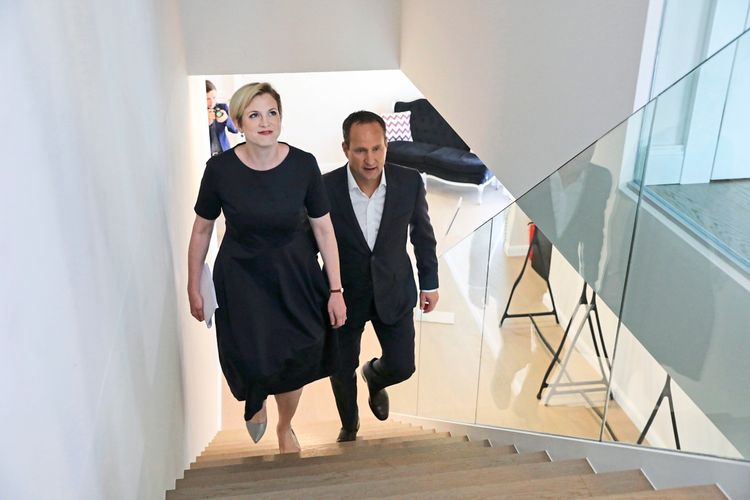 Zwei Personen gehen eine helle, moderne Treppe hinauf. Im Hintergrund sind Möbel und eine weitere Person mit Kamera zu sehen.