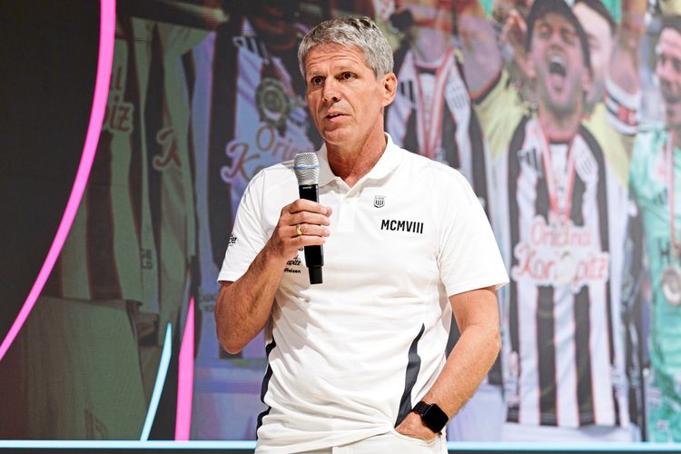 Ein Mann in weißem Polohemd, der ein Mikrofon hält, spricht bei einer Veranstaltung. Im Hintergrund sind jubelnde Personen in Fußballtrikots zu sehen.