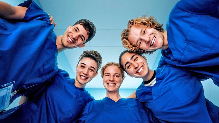 Fünf junge Personen tragen blaue Krankenhauskleidung und stehen im Kreis, während sie glücklich in die Kamera lächeln. Der Hintergrund zeigt eine helle Krankenhausumgebung.
