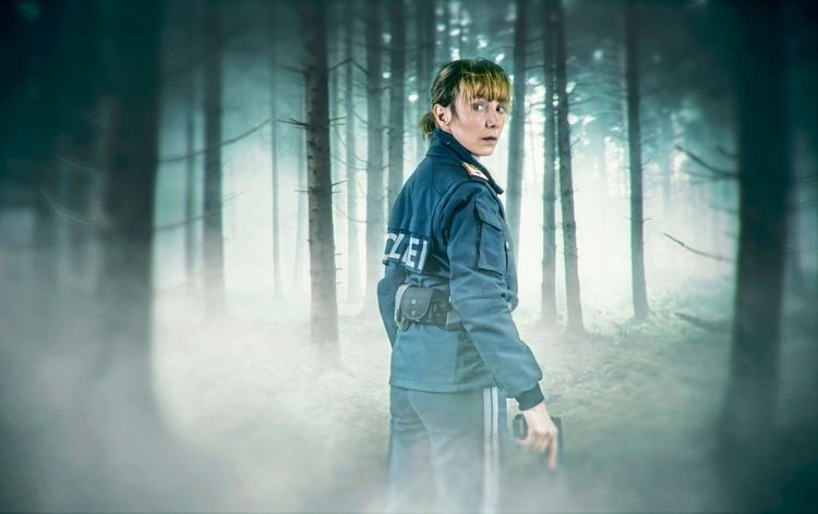 Eine Polizistin in blauer Uniform mit der Aufschrift „Polizei“, bewaffnet und besorgt dreinschauend, steht in einem nebligen Wald. Die Szene wirkt düster und geheimnisvoll.
