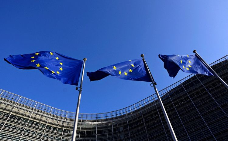 Drei Europaflaggen mit gelben Sternen auf blauem Hintergrund wehen vor dem Hauptgebäude der Europäischen Kommission in Brüssel, Belgien, unter klarem blauem Himmel.
