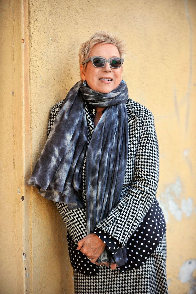 Doris Dörrie steht mit Sonnenbrille, weitem Schal und Mantel an eine gelbe Hausmauer gelehnt.