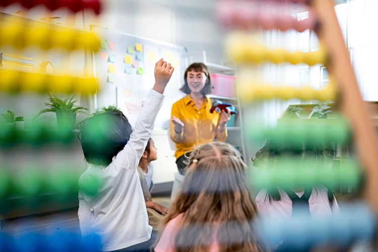 In einem Klassenzimmer sitzt eine Gruppe von Kindern auf dem Boden und hört aufmerksam einer Lehrperson zu, die ein Spielzeugflugzeug in der Hand hält. Im Vordergrund ist ein großer Abakus sichtbar, der das Bild teilweise verdeckt. Im Hintergrund hängen bunte Haftnotizen an einer Tafel.