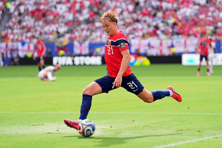 Ein Fußballspieler der norwegischen Nationalmannschaft in rotem Trikot und blauer Hose schießt während eines Spiels auf das Tor. Im Hintergrund sind Zuschauer und eine Spielfeldbande mit der Aufschrift 