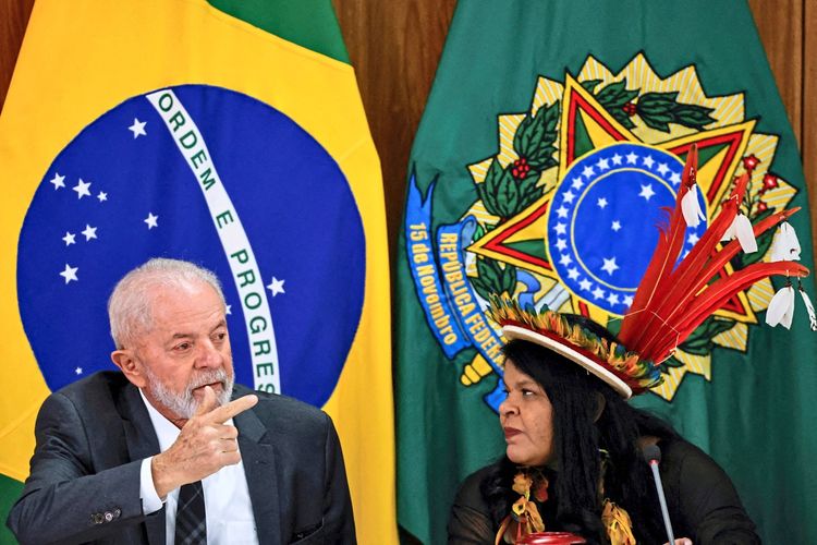 Ein Mann in Anzug und Krawatte und eine Frau in traditioneller indigener Kleidung mit Federschmuck sprechen am Tisch miteinander. Im Hintergrund sind zwei brasilianische Flaggen zu sehen.