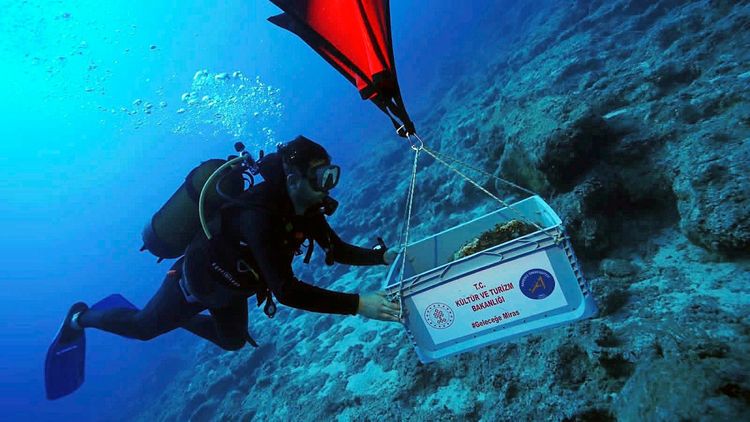 Ein Taucher unter Wasser hebt mit Hilfe eines Hebesystems eine Kiste, die mit einer Aufschrift des türkischen Kultur- und Tourismusministeriums versehen ist, vom Meeresboden an die Oberfläche.