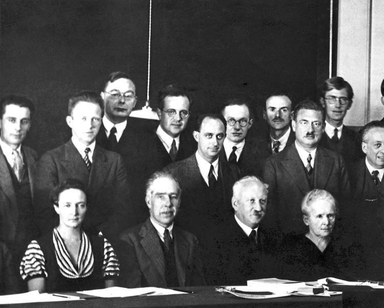 Schwarz-Weiß-Fotografie einer Gruppe von Physikern bei der siebten Solvay-Physikkonferenz in Brüssel, Belgien, Oktober 1933. Im Vordergrund sitzen (von links nach rechts): Irène Joliot-Curie, Niels Bohr, Abram Ioffe und Marie Curie. Dahinter stehen mehrere Personen, darunter identifizierbare Wissenschaftler wie Werner Heisenberg, Hendrik Kramers, Enrico Fermi und andere. Im Hintergrund eine dunkle Wand und eine herabhängende Lampe.