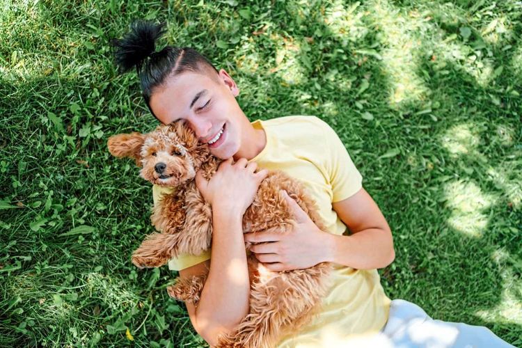 Person mit kurzen schwarzen Haaren liegt auf dem Rücken im Gras und kuschelt mit einem flauschigen Hund