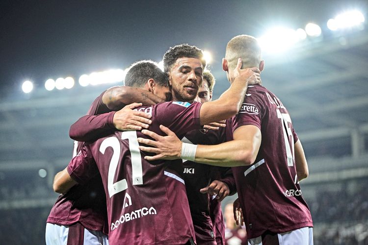 Eine Gruppe von Fußballspielern des Torino FC feiert gemeinsam ein Tor im Stadion, alle tragen bordeauxrote Trikots. Hintergrund von Flutlicht beleuchtet.
