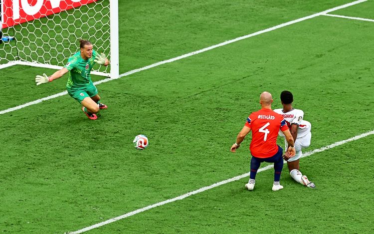 Ein Fußballspiel während des FIFA-Weltmeisterschafts-2026-Viertelfinales zwischen Norwegen und England. Jude Bellingham von England erzielt ein Tor gegen den norwegischen Torhüter Orjan Nyland. Ein norwegischer Verteidiger ist ebenfalls in der Szene zu sehen. Im Hintergrund das Tor und der grüne Rasen.