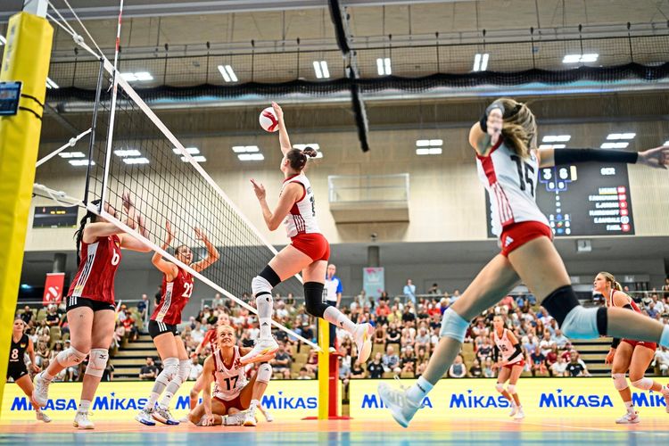 Eine Spielsituation während eines Volleyball-Testspiels der Frauen zwischen Österreich und Deutschland am 15. August 2026 in Wien. Eine Spielerin in Rot „blockt“ nah am Netz, während andere Spielerinnen in Aktion sind. Zuschauer und eine Anzeigetafel im Hintergrund.