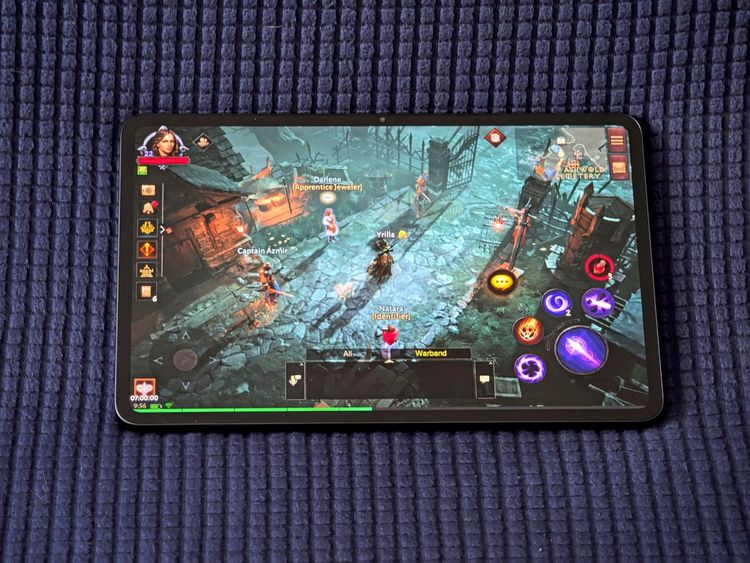 Ein Tablet mit einem Videospiel auf dem Bildschirm. Das Spiel zeigt eine mittelalterliche Fantasyszene mit Figuren in einer gepflasterten Umgebung, umgeben von Gebäuden und Toren. Steuerungssymbole und Spielerinteraktionen sind sichtbar. Das Tablet liegt auf einem blauen texturierten Untergrund.