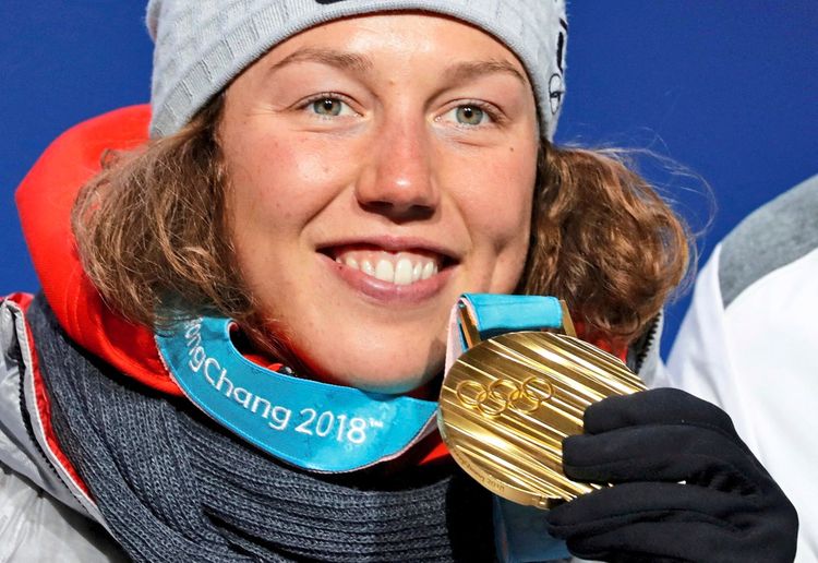 Eine Person trägt eine graue Mütze und hält eine olympische Goldmedaille mit dem Schriftzug „PyeongChang 2018“. Die Medaille hängt an einem türkisfarbenen Band, die Person lächelt.