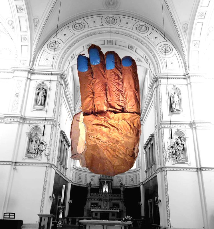 Im Altarraum der Breitenfelder Kirche in Wien hängt eine überdimensionierte Hand, die aus Stoff genäht ist, die Hautfarbe ist dunkler, die Fingernägel sind blau lackiert. Der Kirchenhintergrund ist in Schwarzweiß. 