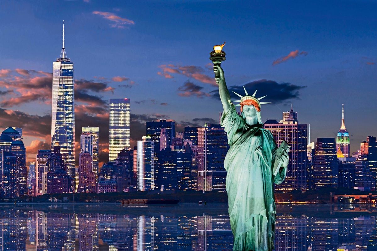 Die Freiheitsstatue im Vordergrund vor der Skyline von New York City bei Sonnenuntergang. Links ist das neue World Trade Center zu sehen, rechts das Empire State Building. Die Gebäude und die Lichter spiegeln sich im Wasser.