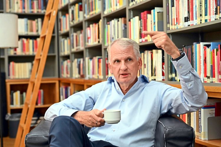 Ein Mann sitzt in einem Raum mit Bücherregalen im Hintergrund. Er trägt ein hellblaues Hemd, hält eine Tasse in der Hand und gestikuliert mit der anderen Hand.