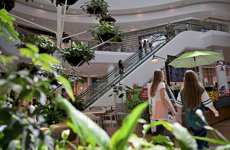 In einem modernen Einkaufszentrum mit mehreren Stockwerken und viel Grün sind Menschen auf Rolltreppen und in einem Café-Bereich zu sehen. Im Vordergrund befinden sich Pflanzen, während im Hintergrund Geschäfte und ein Stand mit einem grünen Sonnenschirm sichtbar sind.