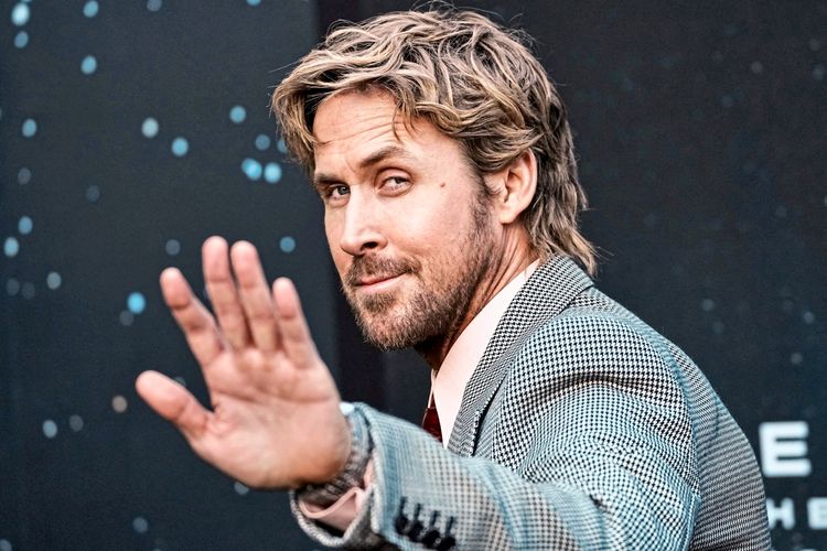 Ein Mann mit blonden Haaren und Bart trägt einen karierten Anzug und hebt eine Hand in einer abwehrenden oder winkenden Geste. Der Hintergrund ist dunkel mit dezenten Lichteffekten.