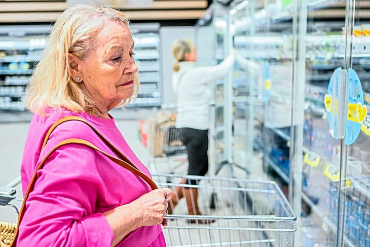 Eine ältere Frau betrachtet nachdenklich die Tiefkühlware in einem Supermarkt.