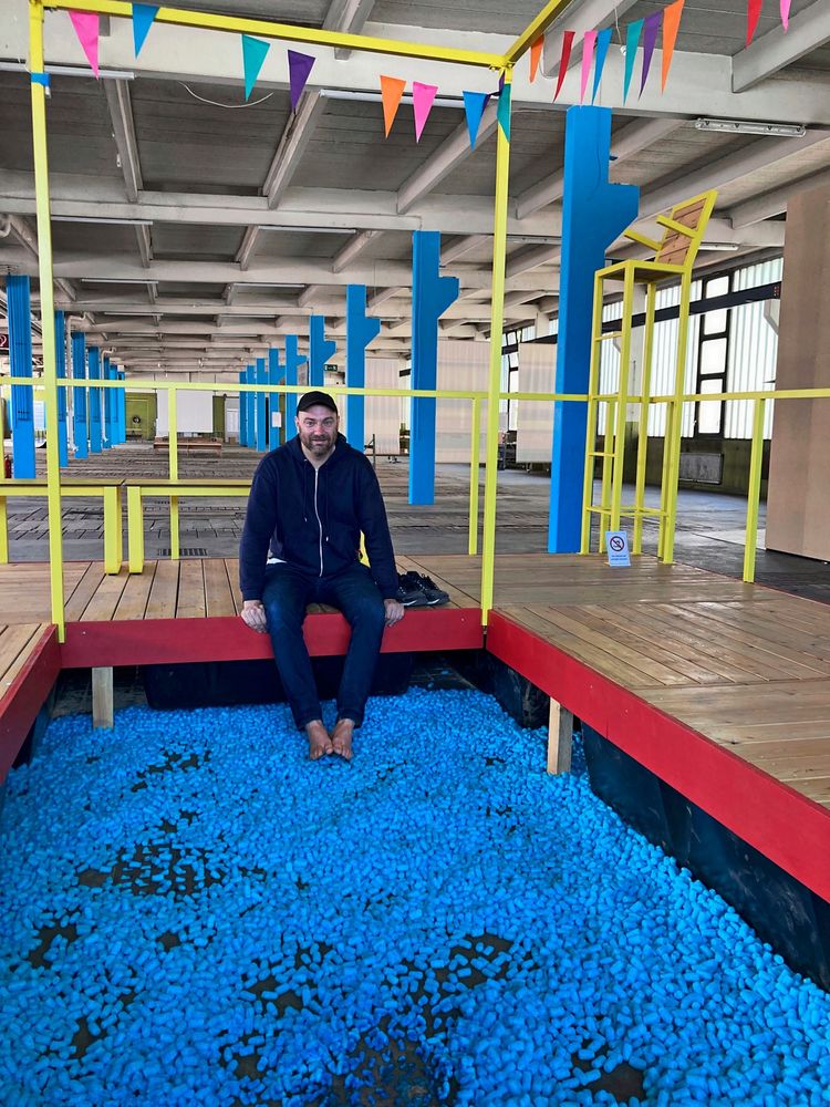 Ein Mann sitzt auf einer hölzernen Plattform und hat seine Schuhe ausgezogen. Unter ihm befindet sich eine Fläche mit blauen Schaumstoffstücken. Die Konstruktion ist von gelben und blauen Streben umgeben, und oben hängen bunte Wimpel. Der Hintergrund zeigt eine weitläufige, industrielle Halle.
